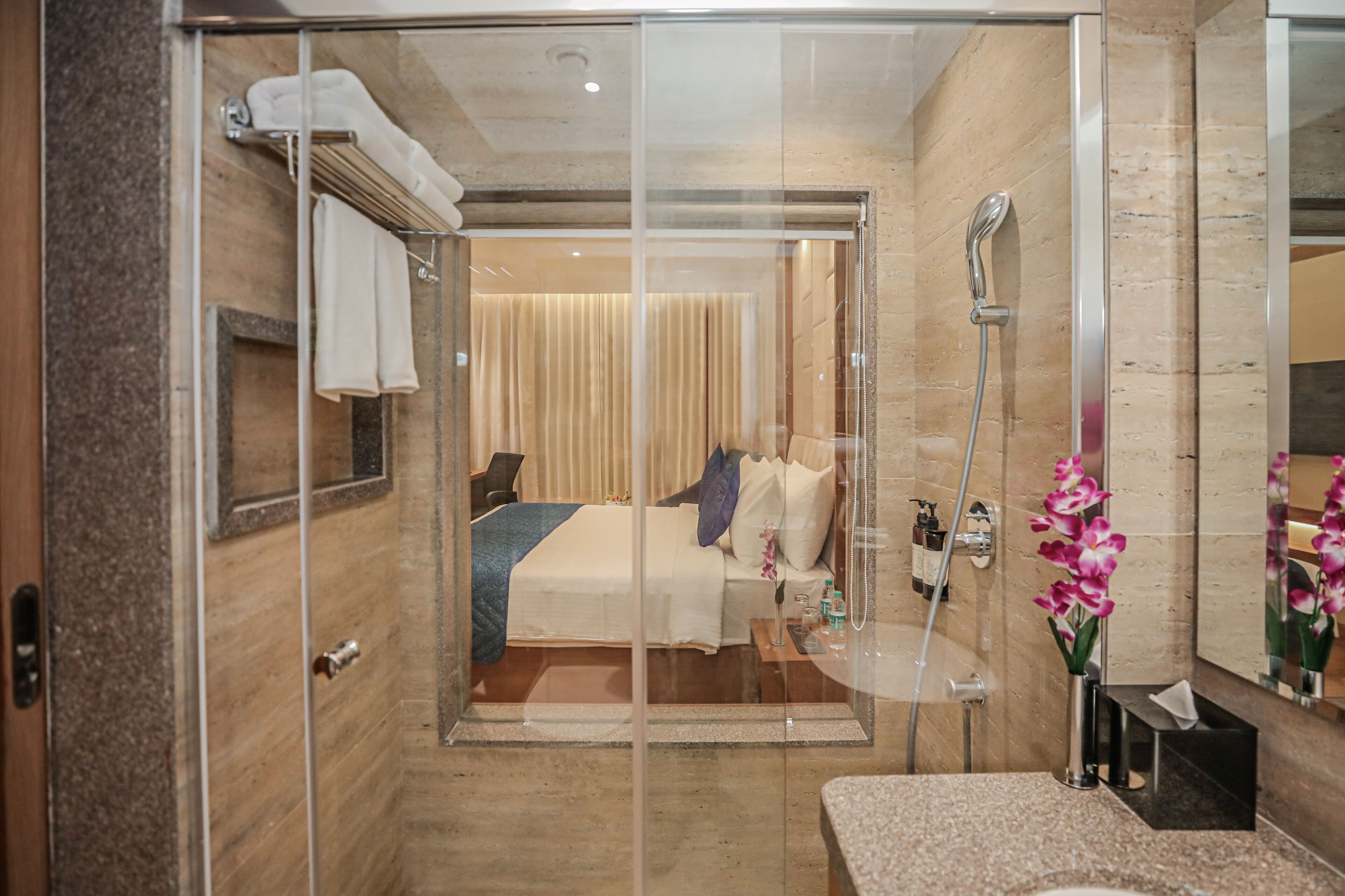 A hotel room with a glass shower enclosure, a bed with a blue blanket, a bedside table with toiletries, and a large mirror.