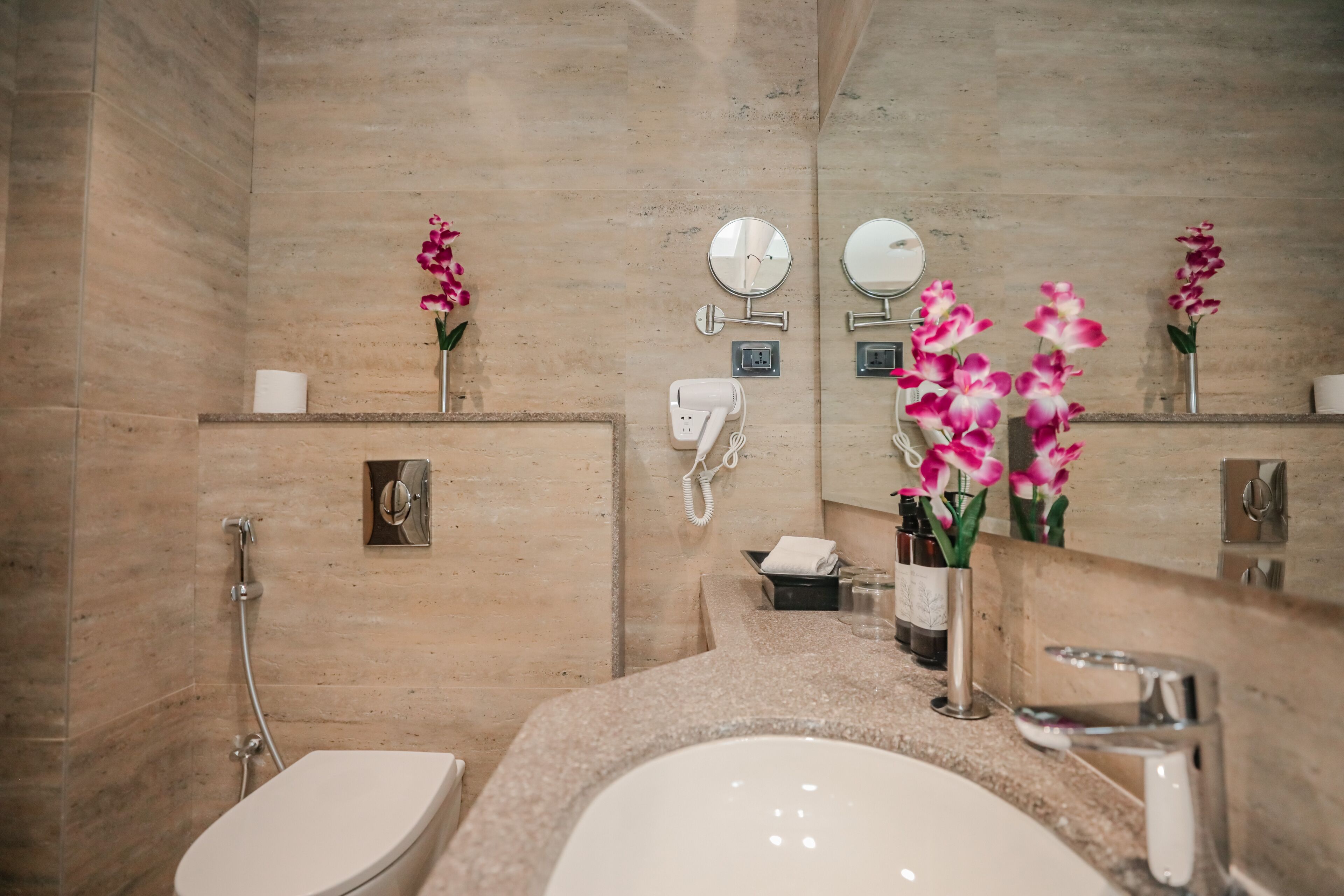 A modern bathroom with a beige marble wall, a white sink, a mirror, and two orchid vases.