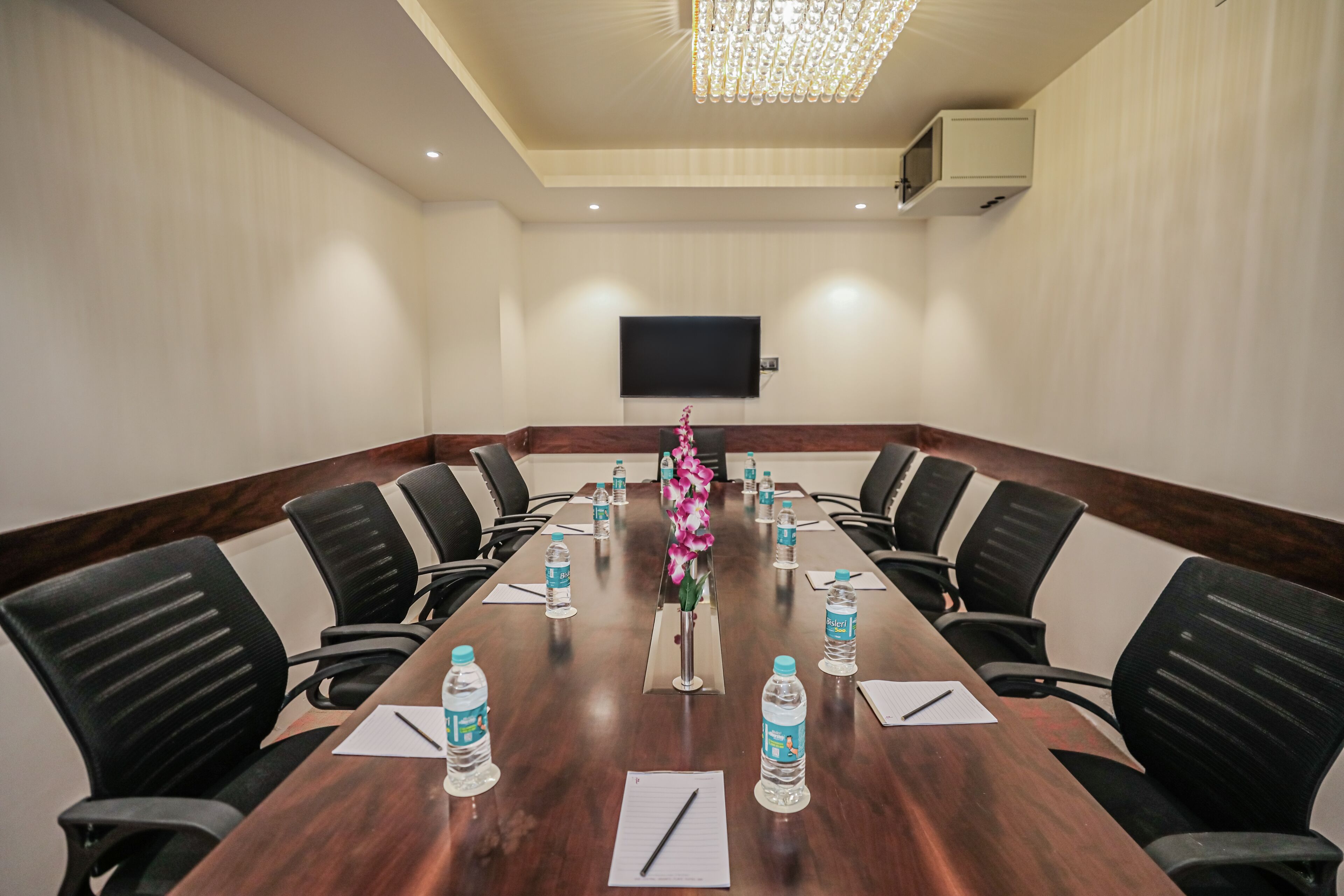 A conference room with a long table, black chairs, water bottles, a flower vase, and a flat-screen TV on the wall.