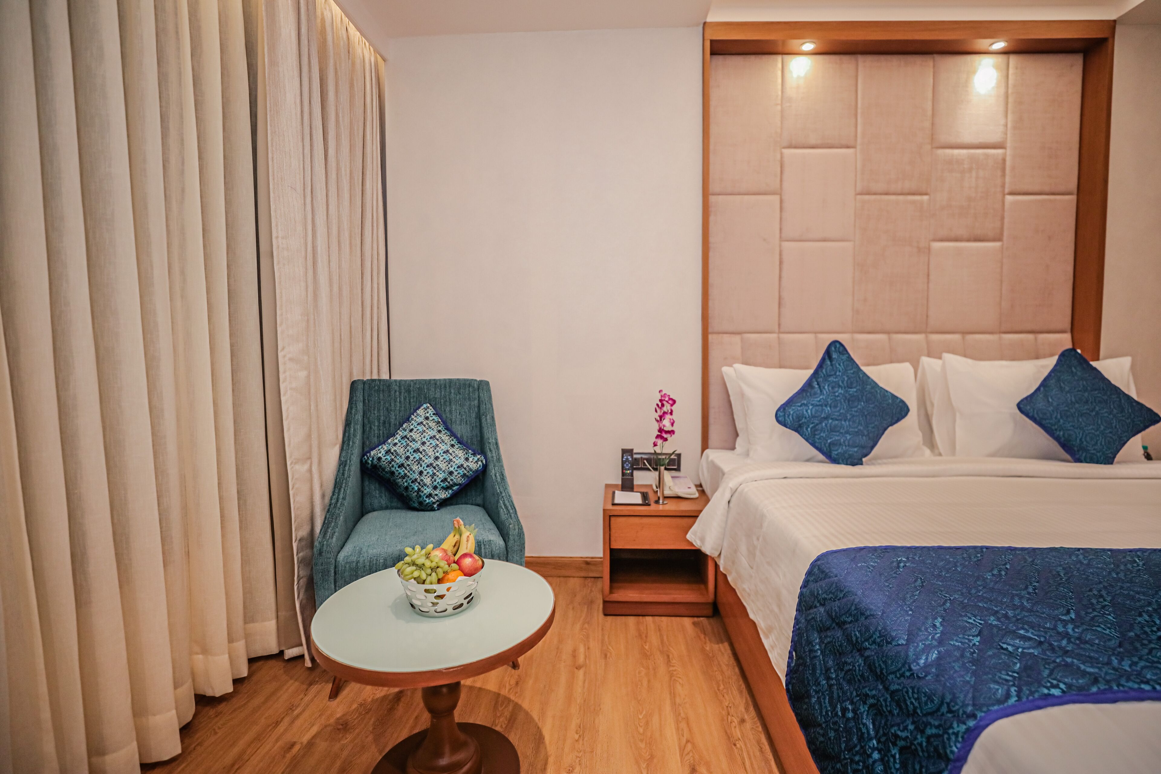 A hotel room with a bed, a chair, a small table with fruit, and a nightstand with a lamp.