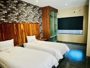 A hotel room with two beds, a brick-patterned wall, a wooden headboard, a green shelving unit, and a window with a blind. - The Outlook At Victoria Falls (Victoria Falls)