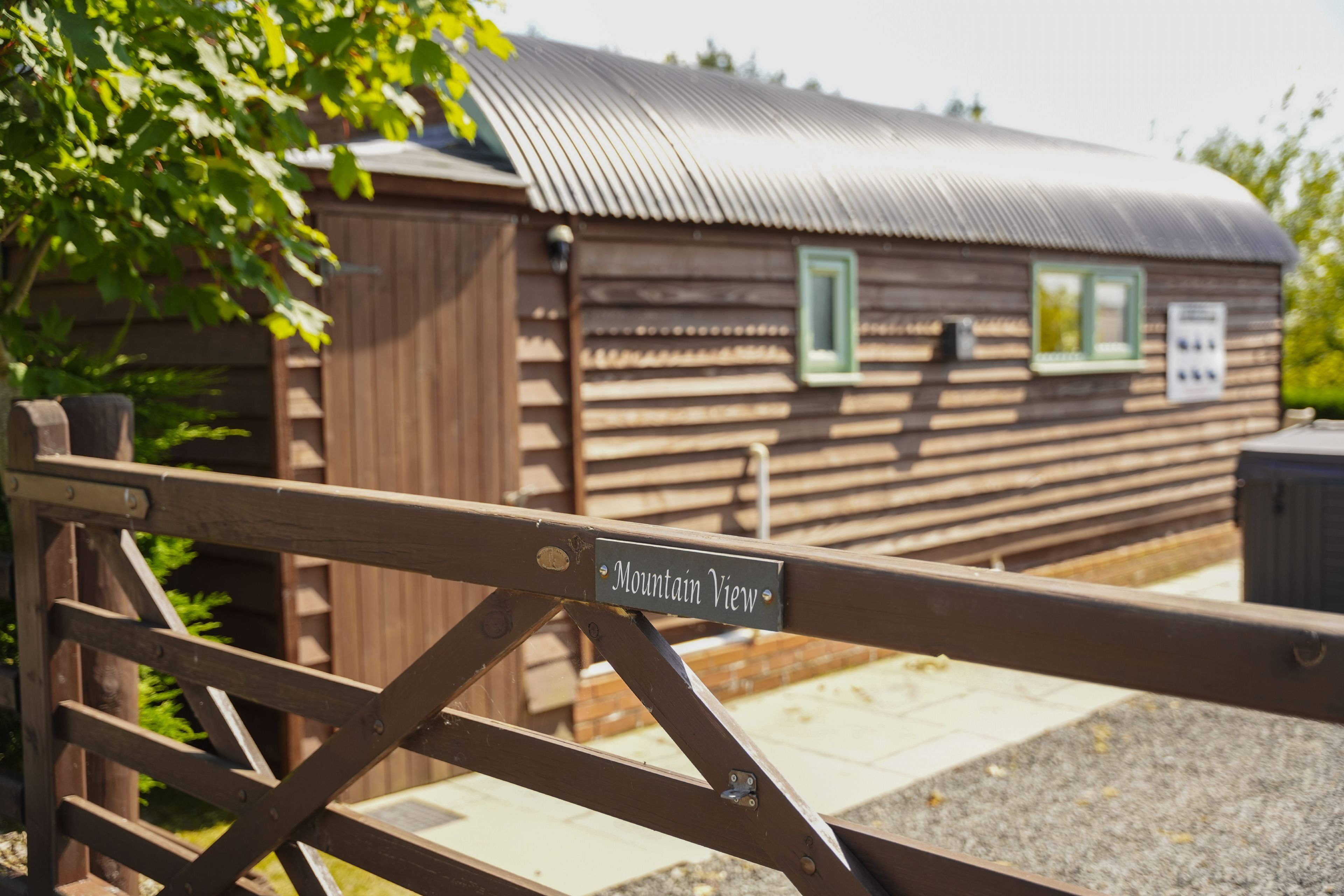 A wooden cabin with a metal roof, a wooden gate with a sign reading 'Mountain View,' and a brick pathway leading to the cabin.