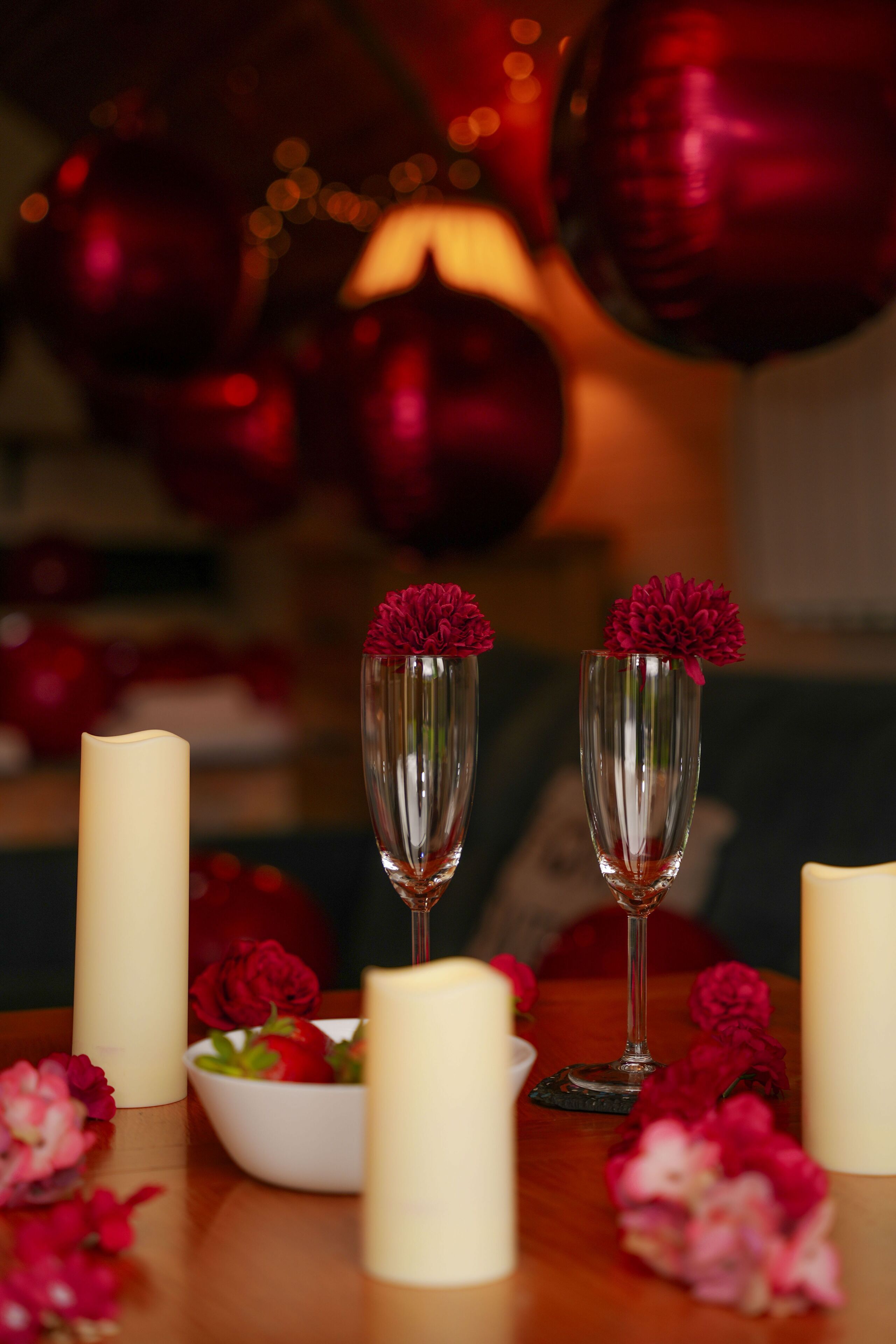 A table with candles, a bowl of flowers, and two champagne flutes with red flowers.