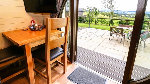 Een houten interieur met een televisie, een tafel en een stoel. Buiten is er een terras met een tafel en stoelen, en uitzicht op groen en heuvels.