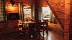 L’intérieur chaleureux d’un chalet avec une table et des chaises en bois, des rideaux à carreaux, une cheminée et de grandes fenêtres donnant sur la forêt.