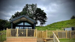 A small, modern building with a wooden deck and a glass entrance, surrounded by a wooden fence and a grassy hill.