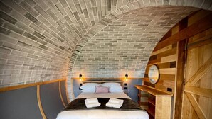Une chambre confortable avec un plafond en briques incurvé, des poutres en bois, un lit avec des oreillers et des serviettes, et une table de chevet en bois.