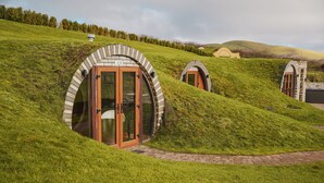 Une série de structures circulaires, recouvertes d’herbe, dotées de portes en bois et de murs en briques, situées sur une colline verdoyante.