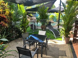 A patio area with a table and chairs, surrounded by various plants and a pool in the background.