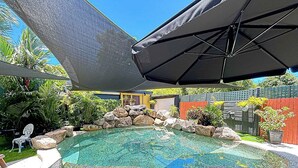 Una piscina con diseño de guijarros, rodeada de rocas y plantas, con una zona sombreada.