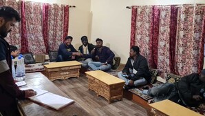 A group of people seated in a room with patterned curtains, wooden furniture, and a table with a notebook and water bottle.