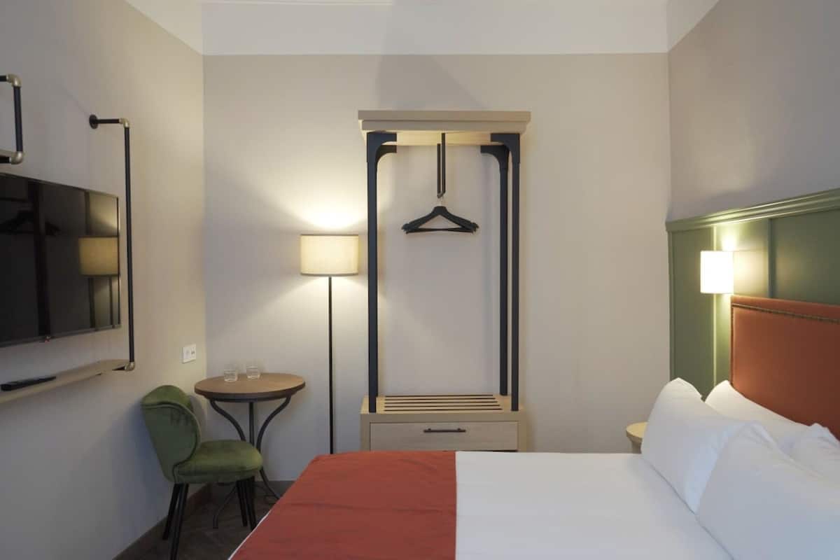 A hotel room with a bed, a chair, a small table, a lamp, a TV, and a coat rack.