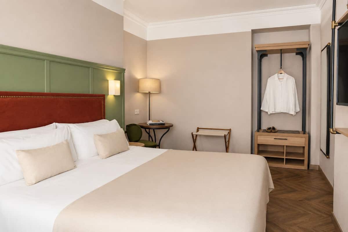 A hotel room with a bed, a bedside table, a chair, a lamp, a coat rack, and a mirror.