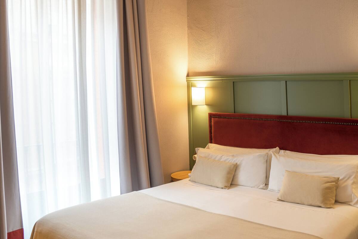 A hotel room with a bed, pillows, a nightstand, and a wall-mounted lamp.