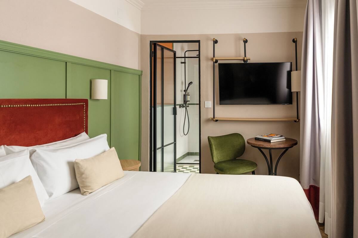 A hotel room with a bed, a green chair, a small table, a TV, and a shower area.