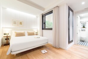 Habitación de hotel moderna con una cama grande, una ventana con cortinas, una mesita de noche con lámpara y vistas al baño.