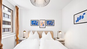 Une chambre d’hôtel avec un lit, deux lampes de chevet, une fenêtre avec des rideaux et trois œuvres d’art encadrées accrochées au mur.