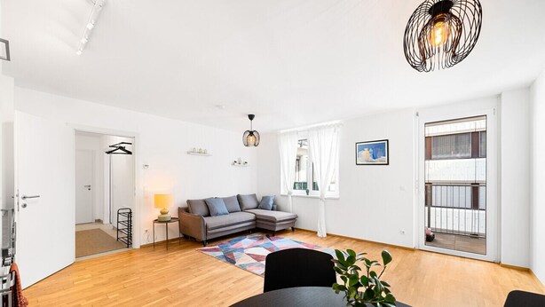 Un salon moderne avec un canapé d’angle gris, une table de salle à manger ronde et une grande fenêtre donnant sur un balcon.