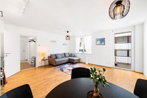 Un salon moderne avec un canapé d’angle gris, une table de salle à manger ronde et une grande fenêtre donnant sur un balcon.