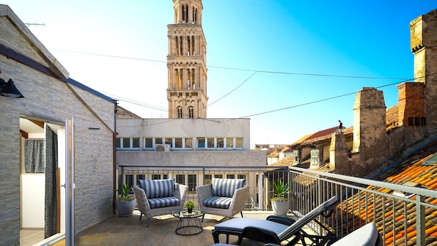 A balcony with wicker furniture, a view of a tower, and a building with a tiled roof. - Sirius Luxury Rooms (Split)