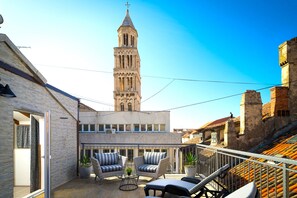 Un balcón con mobiliario de mimbre, vista a una torre y un edificio con tejado de tejas.