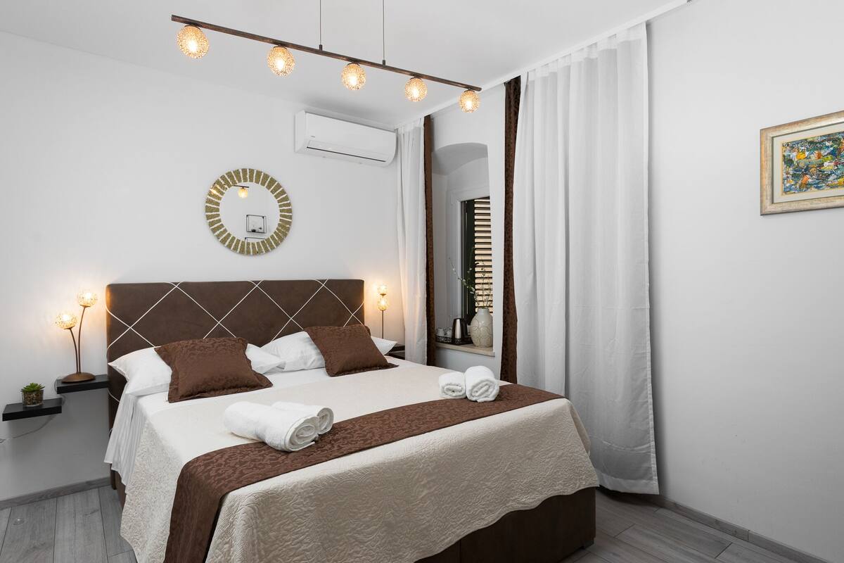Una cama bien hecha con cabecera marrón, sábanas blancas y dos toallas. Encima de la cama, en la pared, hay un espejo redondo y, al lado, una planta pequeña en la mesita de noche.