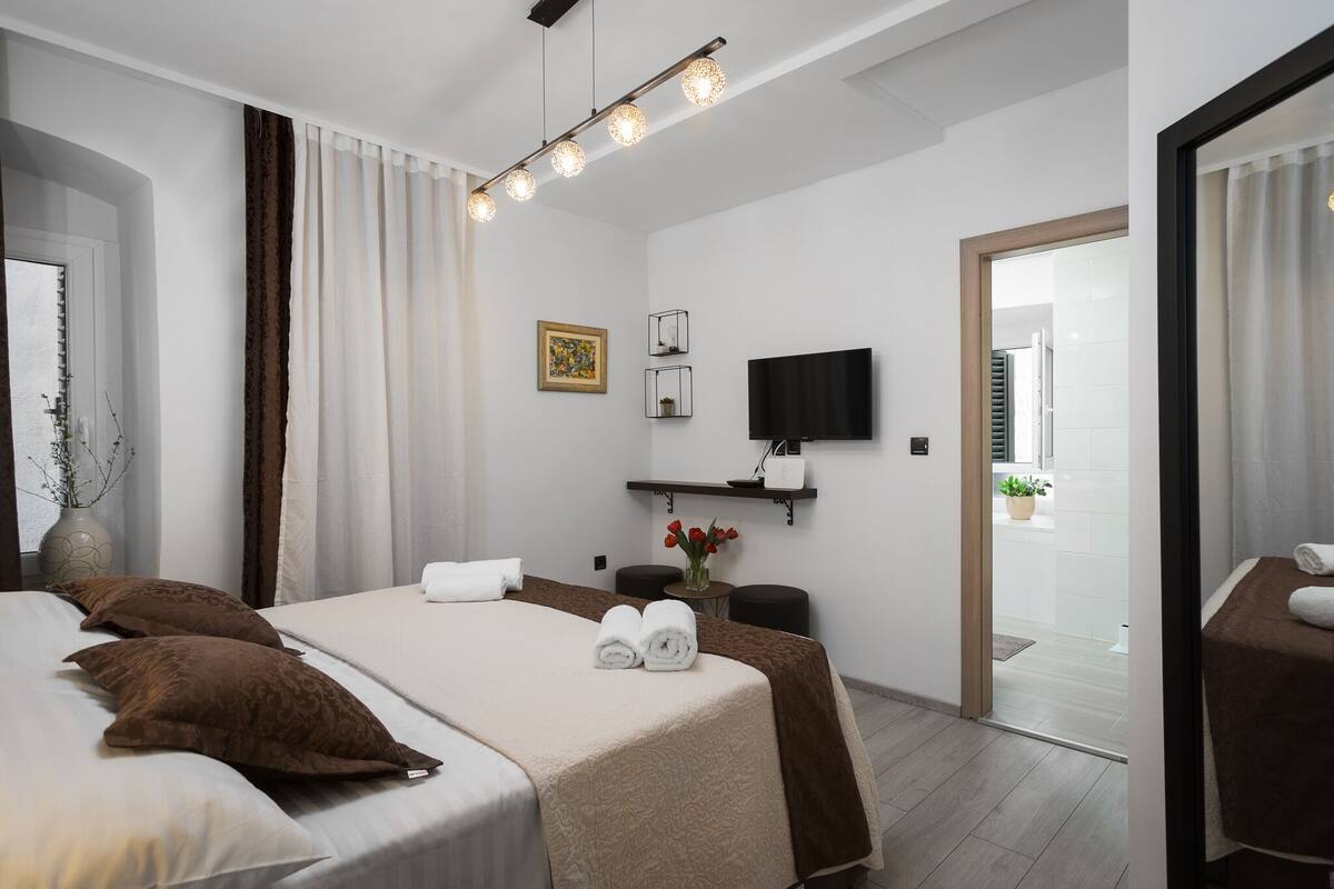 Una habitación de hotel con cama, televisión, una mesita con un jarrón de flores y una puerta que da a un baño.