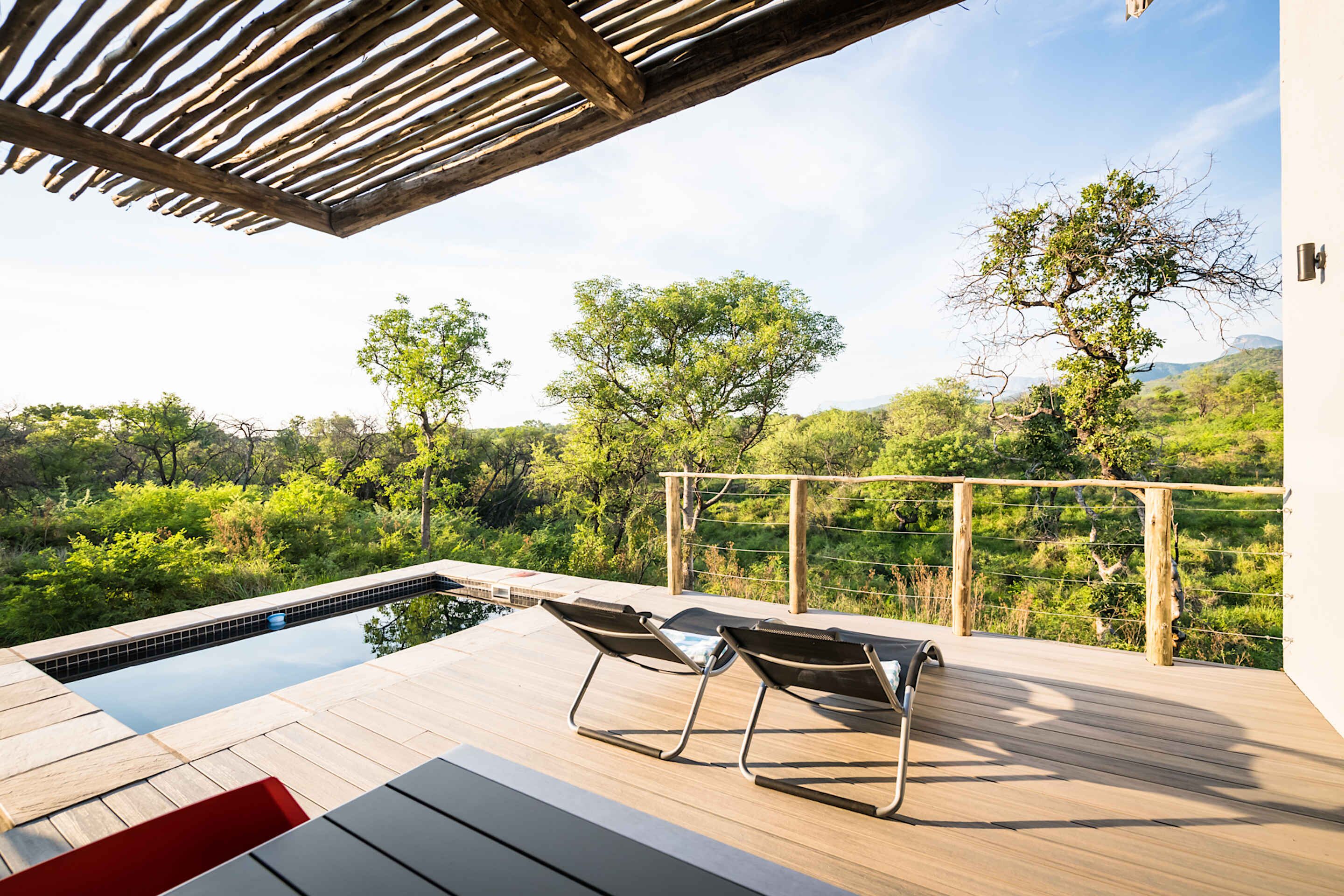A wooden deck with a pool, two lounge chairs, and a view of a lush landscape.