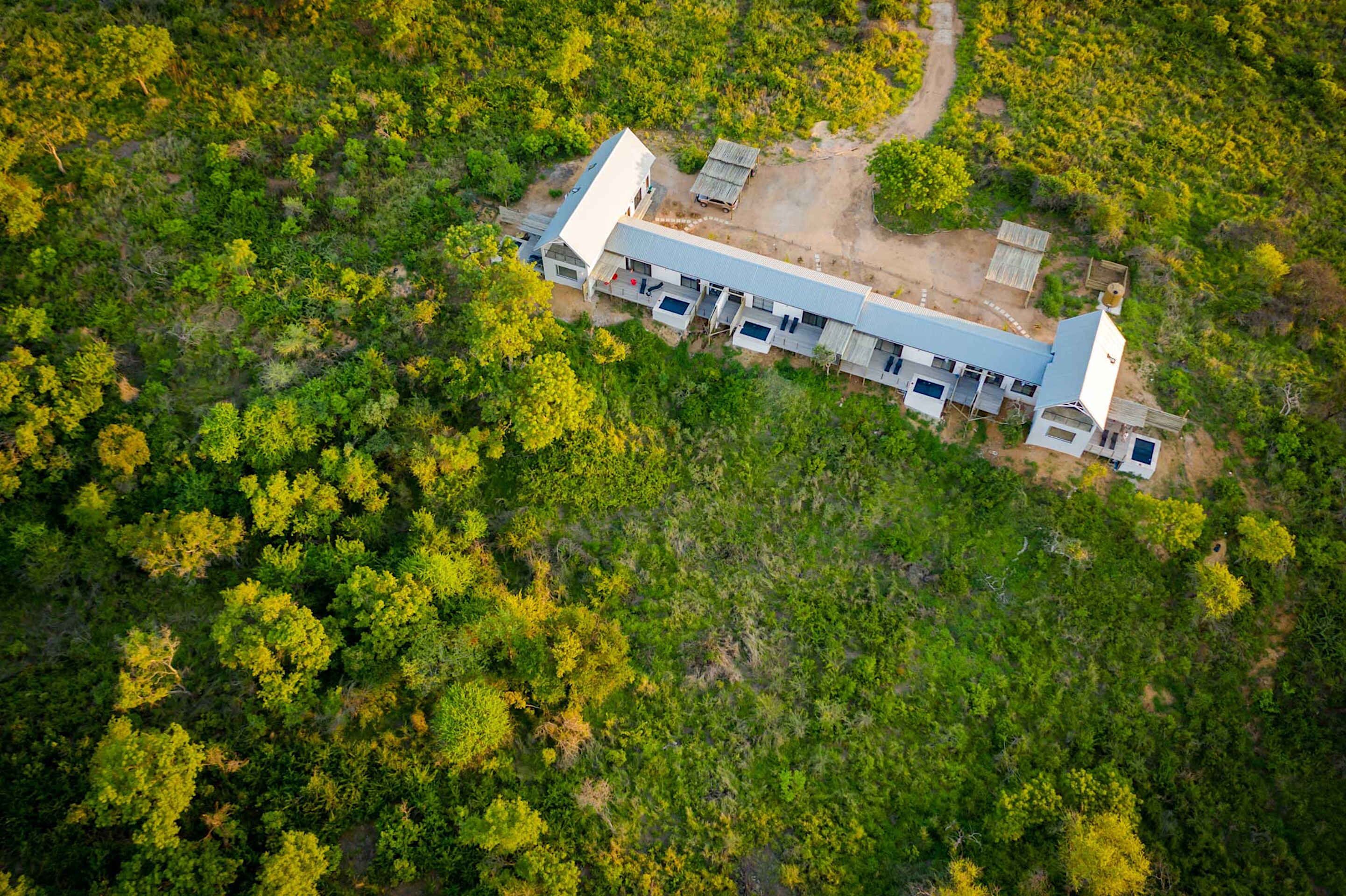An aerial view of a long, narrow building with a flat roof, surrounded by dense green forest.