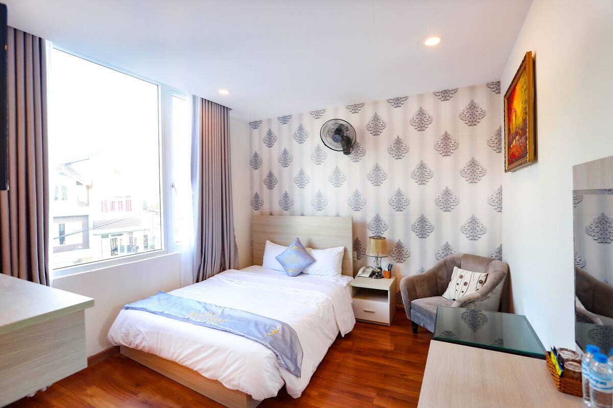 A hotel room with a bed, a chair, a desk, a nightstand, a lamp, and a window with curtains.