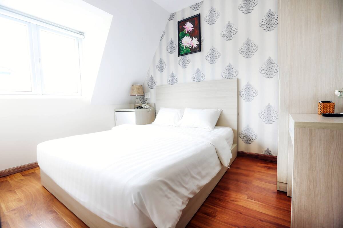 A neatly made bed in a room with a wooden floor, a nightstand with a lamp, and a wall with a floral pattern.