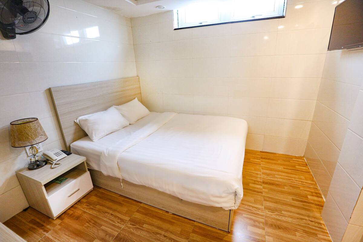 A neatly made bed in a room with a wooden floor, a bedside table with a lamp, and a wall-mounted fan.