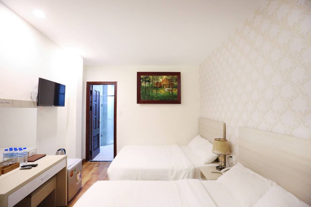 A hotel room with two beds, a desk with a flat-screen TV, a painting on the wall, and a door leading to a balcony.
