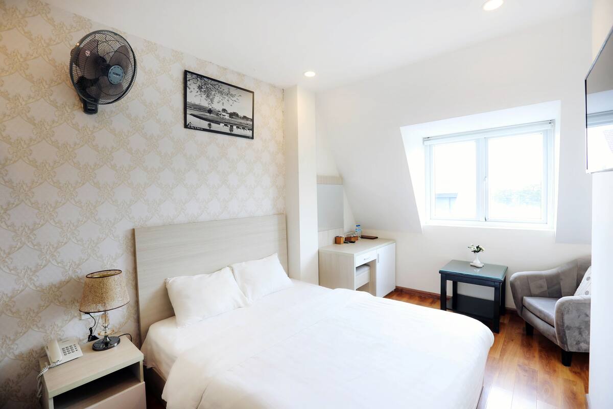 A hotel room with a bed, bedside table, lamp, fan, desk, chair, and a window.
