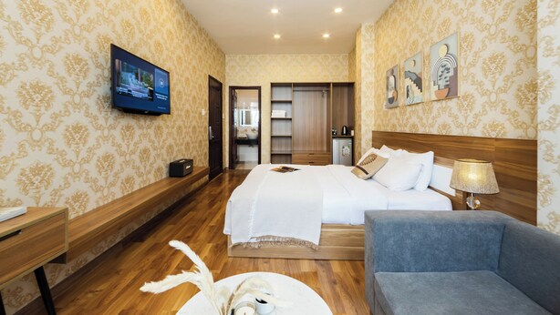 A hotel room with a large bed, a TV mounted on the wall, a wooden bench, a blue armchair, and a small table with a decorative item. - TTR Vincent Hotel (Xuan Huong - Da Lat)