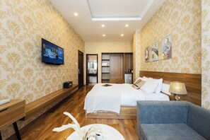 A hotel room with a large bed, a TV mounted on the wall, a wooden bench, a blue armchair, and a small table with a decorative item. - TTR Vincent Hotel (Xuan Huong - Da Lat)