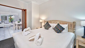 A hotel room with a bed, two pillows, a nightstand, a lamp, a chair, a table, and a view of a balcony with a chair and a plant. - Alassio 2 Bedroom Direct Pool Access104 (Palm Cove)