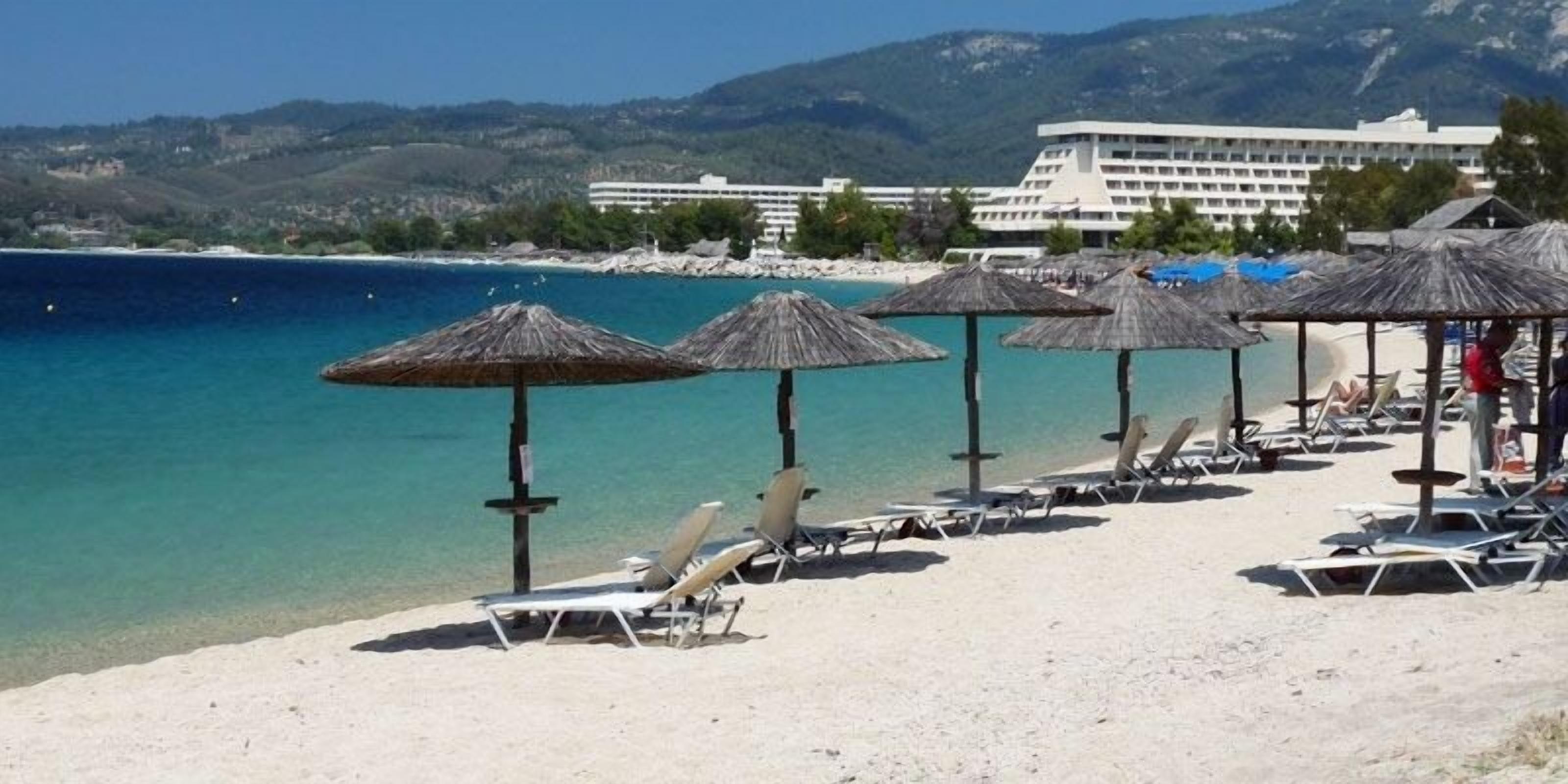 A beach with sun loungers and thatched umbrellas, a clear blue sea, and a hotel in the background.