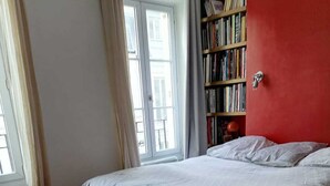 Een slaapkamer met een houten vloer, een bed met wit beddengoed, een boekenkast en een raam met gordijnen.