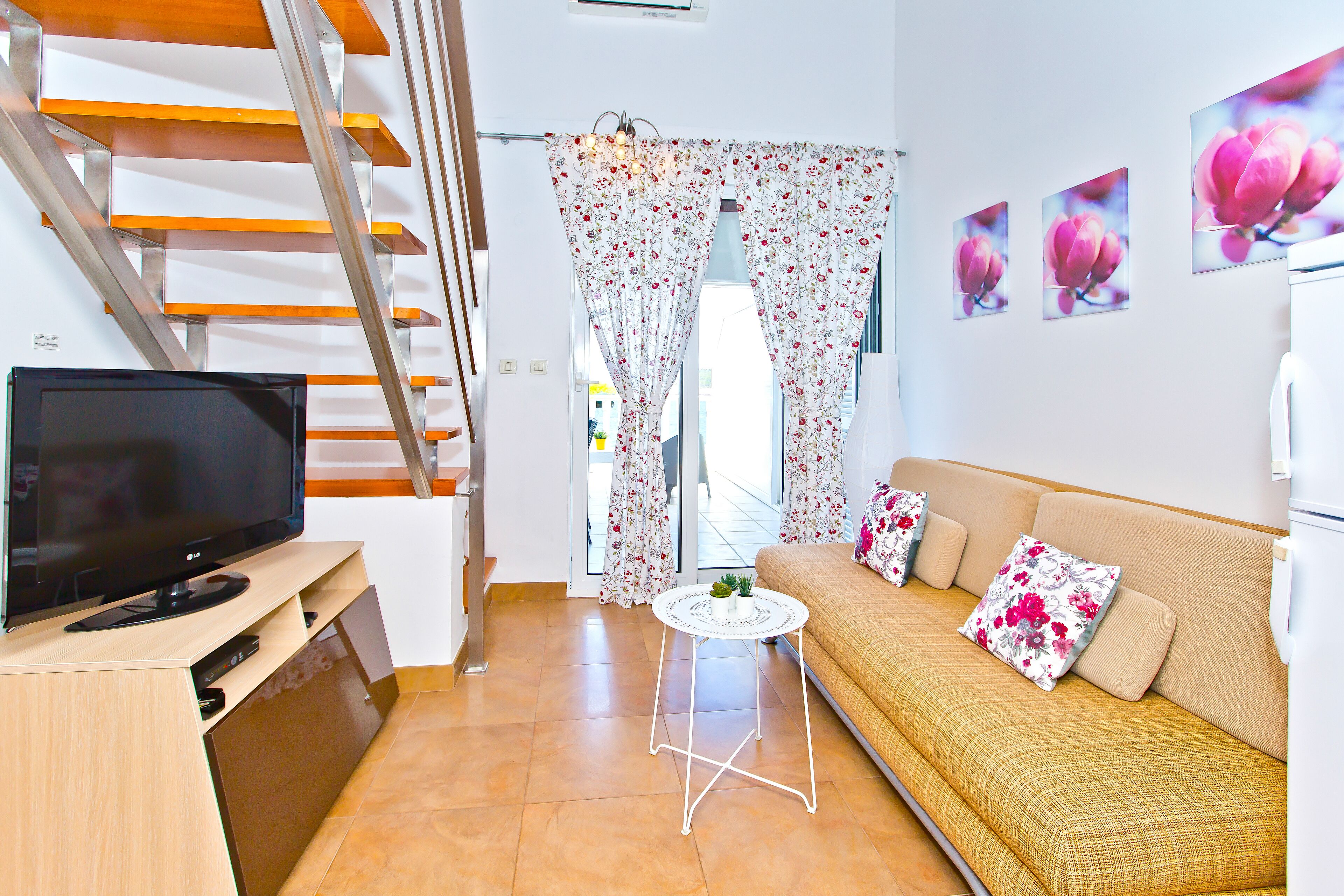 A modern living room with a flat-screen TV, a sofa with floral cushions, a small round table, and a staircase with wooden steps.