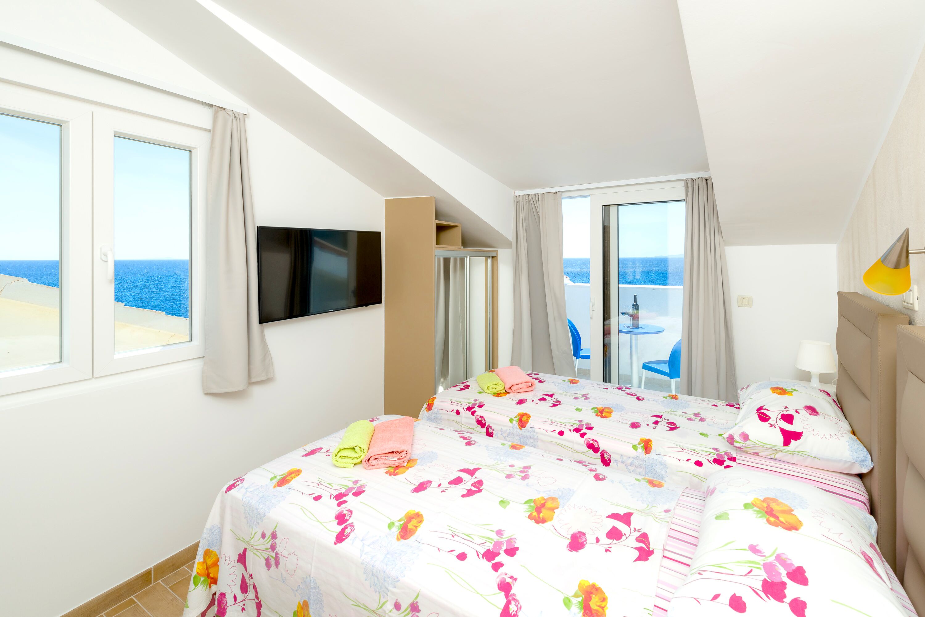 A hotel room with two beds, a TV, a balcony with a view of the sea, and a small table with a chair outside.