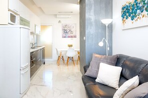 Et moderne køkken med hvide hårde hvidevarer, et spiseområde med et hvidt bord og stole, samt en sort lædersofa med puder.