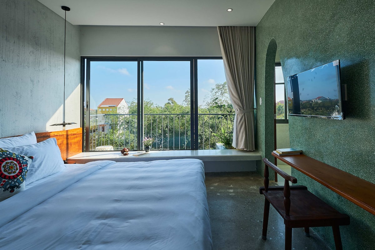 Habitación de hotel con una cama grande, un banco de madera, un televisor montado en la pared y un balcón con vistas a zonas verdes y edificios.