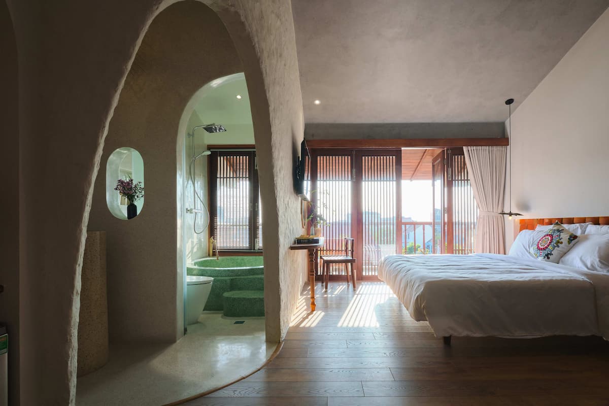 Un dormitorio con una cama grande, piso de madera, vista por una puerta y una pequeña mesa con una silla.