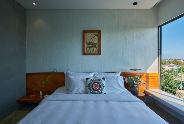 A modern bedroom with a large bed, a wooden headboard, a wall-mounted artwork, a bedside table with a plant, and a view of the city through a large window.