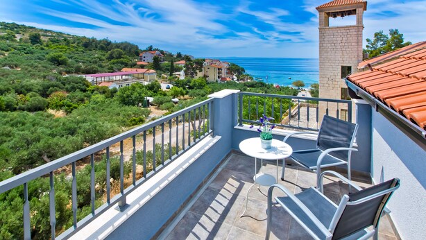 A balcony with a table and chairs, overlooking a coastal area with buildings and trees. - Apartments Maslina (Hvar)