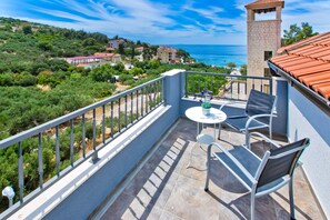 A balcony with a table and chairs, overlooking a coastal area with buildings and trees. - Apartments Maslina (Hvar)