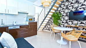 A modern kitchen with white cabinets, a built-in oven, and a coffee maker. There is a dining area with a white table and wicker chairs. A staircase with a black and white leaf pattern leads to another level. - Apartments Maslina (Hvar)