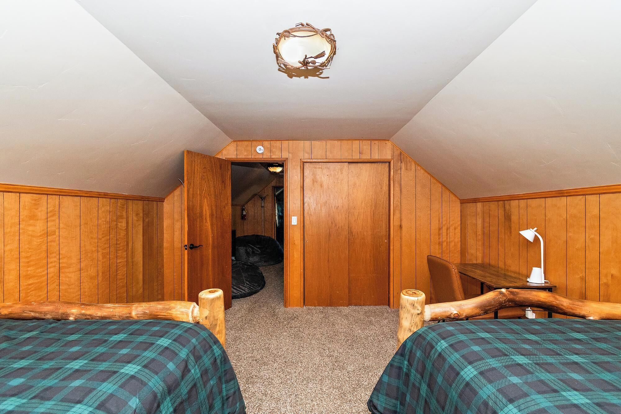 A bedroom with two beds, wooden walls, a ceiling light fixture, and a door leading to another room.