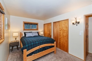 A bedroom with a wooden bed, a nightstand, a lamp, a window with blinds, and a wardrobe. - Golden Bear Pines by Destination Big Bear: Walk to Snow Summit! Fenced Yard! Pet Friendly! HOT TUB! (Big Bear Lake)